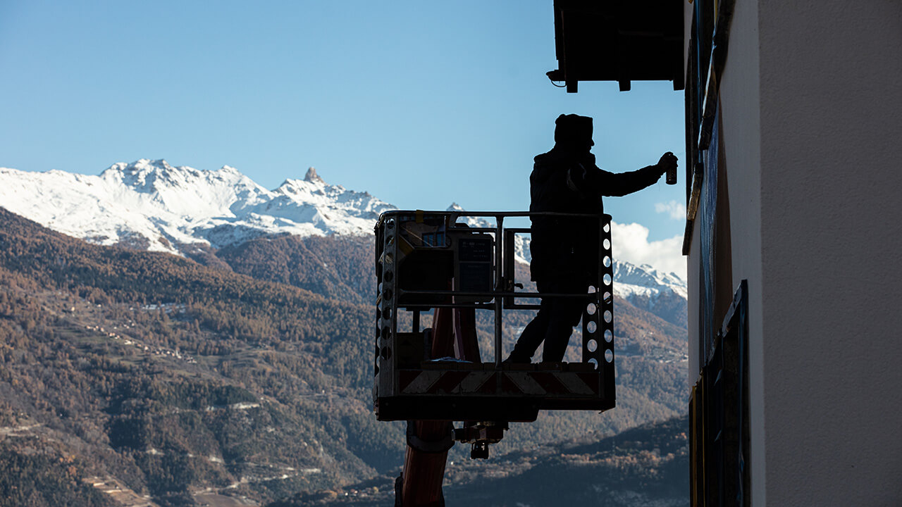 Jasm One peignant une fresque murale en nacelle, Alpes valaisannes en arrière-plan, Art Valais
